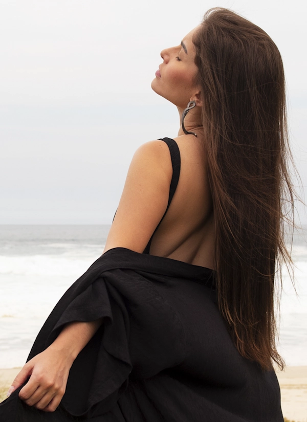 Жінка з довгим волоссям у сукні з відкритою спиною позує на фоні моря, демонструючи гладку спину після пластики